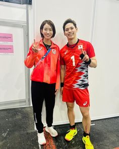 two people standing next to each other in front of a white wall wearing red and black uniforms