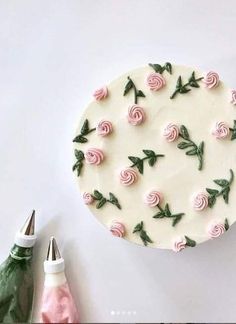 a cake decorated with pink roses and green leaves on a white table next to a pair of crochet hooks