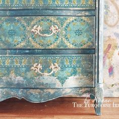 an old dresser painted in blue and green with ornate designs on the top, sitting on a wooden floor