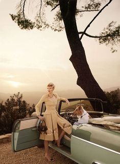 a woman standing next to an old car with her dog sitting on the back seat