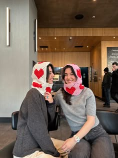 two women wearing knitted hats and scarves are sitting on chairs in a lobby