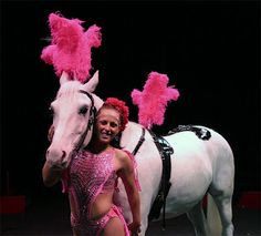 a woman standing next to a white horse with pink feathers on it's head