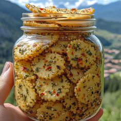 a hand holding a jar full of crackers in front of a mountain range with mountains in the background