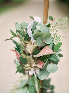 an arrangement of greenery is displayed in a vase on the ground outside, with a wooden stick sticking out of it