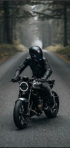 a man riding on the back of a black motorcycle down a road in front of trees