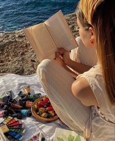 a woman sitting on the beach reading a book next to some crayons and water