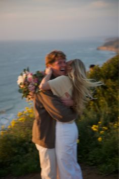 a man and woman hugging each other on the side of a hill near the ocean