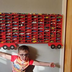 a young boy standing in front of a wall with toy cars on it's back