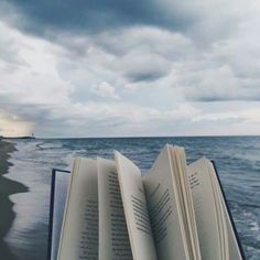 an open book sitting on top of a beach next to the ocean under a cloudy sky