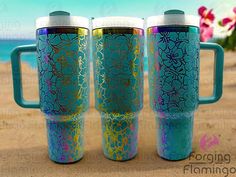 three blue and gold travel mugs sitting on the sand at the beach with flowers in the background