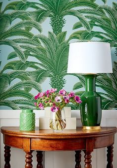 a green vase with pink flowers on a wooden table in front of a palm tree wallpaper