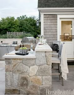 an outdoor kitchen is built into the side of a house with stone counter tops and chairs