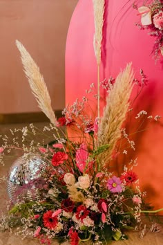 an arrangement of flowers and grasses in front of a disco ball on the floor next to a pink wall