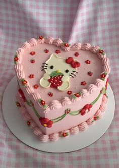 a heart shaped cake with a hello kitty decoration on the top and sides, sitting on a pink checkered tablecloth