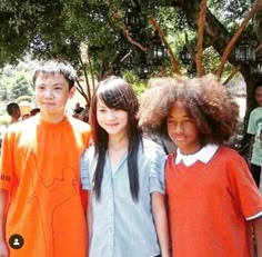 three young children standing next to each other in front of some trees and people looking at the camera