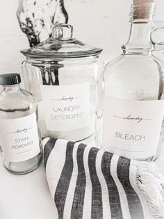 three bottles with labels on them sitting next to a towel and a glass jar filled with liquid