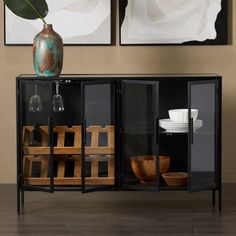 a black cabinet with wine glasses and plates on it in front of two framed paintings