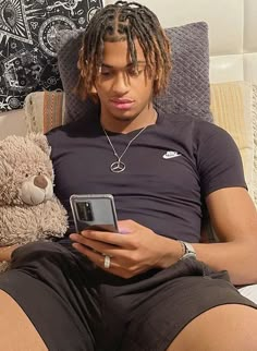 a man with dreadlocks sitting on a couch looking at his cell phone and holding a teddy bear