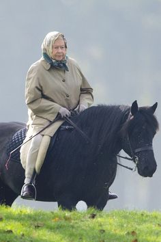 an older woman riding on the back of a black horse in a grassy field with trees behind her