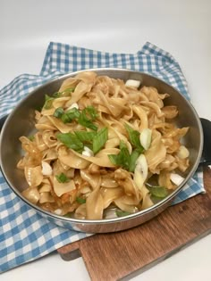 a pan filled with pasta and vegetables on top of a blue checkered cloth next to a wooden cutting board