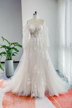 a white wedding dress on display in front of a potted plant and curtained window