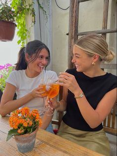 two women sitting at an outdoor table drinking wine and flowers in the vases behind them