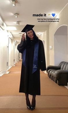 a woman in a graduation gown and cap waves to the camera while standing in a hallway