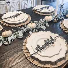 the table is set for thanksgiving dinner with white plates and pumpkins on it, along with greenery