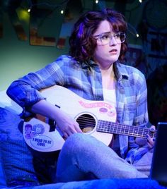 a woman sitting on a bed with a guitar in front of her and a laptop behind her