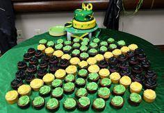 a green table topped with lots of cupcakes next to a yellow and black cake