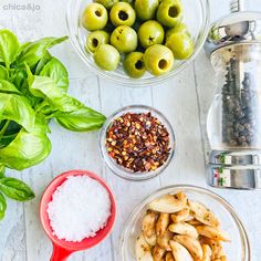 bowls filled with olives, salt and seasoning next to basil leaves on a white table