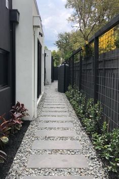 a stone walkway between two buildings with black fence and plants in the foreground on either side