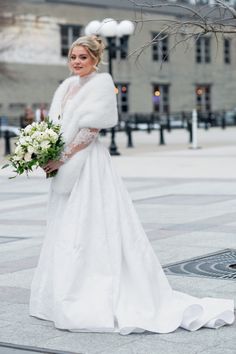 a woman in a white wedding dress and fur stole holding a bouquet on the street