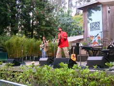 two men are playing music on stage in front of a building with trees and bushes