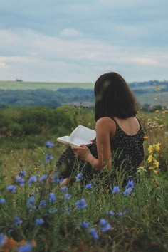 a woman sitting in the grass reading a book with wildflowers around her and hills in the background