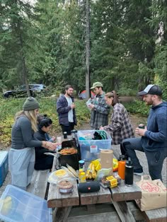 a group of people standing around a picnic table with food on it in the woods