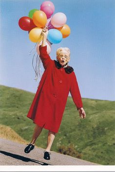 an older woman holding balloons in the air on a road with hills and grass behind her