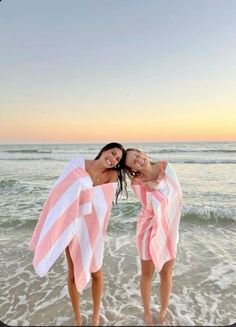 two girls in pink and white towels standing on the beach with their arms around each other