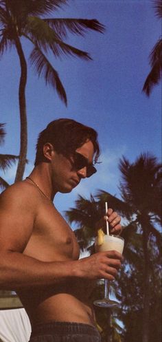 a man in swim trunks holding a drink and looking at his cell phone while standing next to some palm trees