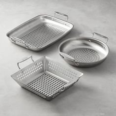 three stainless steel cooking pans with perfored sides and lids on a gray surface