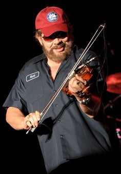 a man with a red hat is holding a violin and looking at the camera while standing on stage