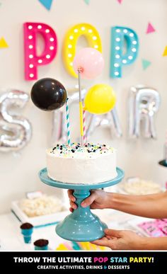 a person holding a cake with candles and balloons on it in front of a party sign