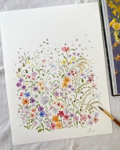 watercolor flowers on paper next to a painting brush and book with colored pencils