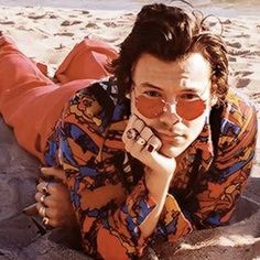 a man laying on the sand with his hands under his chin and wearing sunglasses, looking at the camera