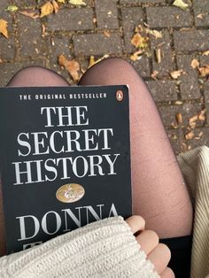 a person holding up a book about the secret history in front of their legs and feet