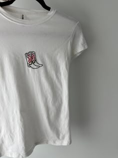 a white t - shirt with a red and black embroidered boot on the front, hanging from a hanger