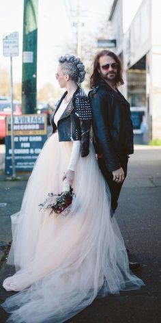 a man and woman are walking down the street in their wedding gowns, one is wearing a black leather jacket