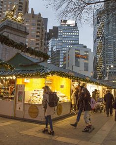 people walking around an outdoor food stand in the city at night with christmas lights on