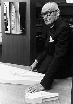 an old man sitting at a table in front of a book case with his hands on it