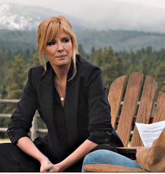 a woman sitting on top of a wooden bench next to a forest filled with trees